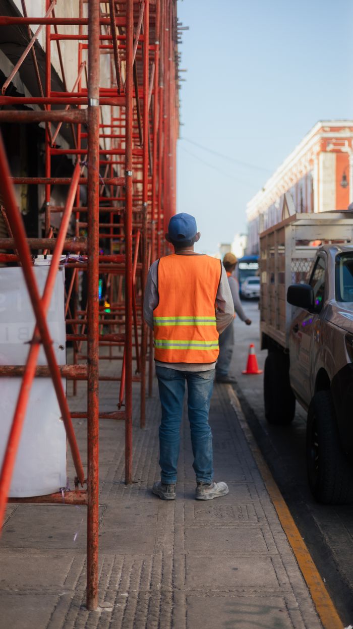 about-01 A construction worker in an orange safety vest stands by scaffolding on a city street.